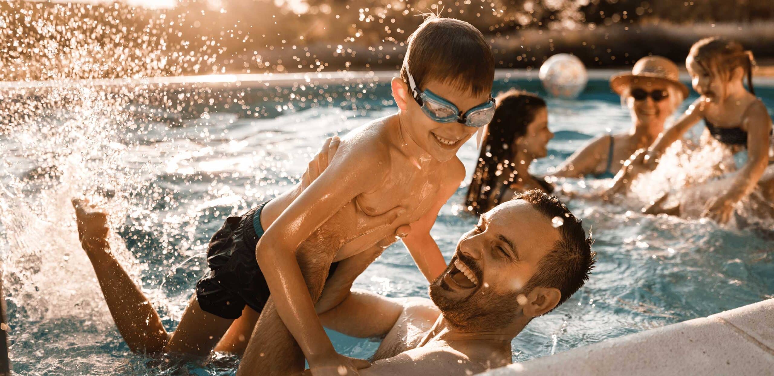 Father holding son with goggles in a backyard pool at sunset, with family playing in the background.