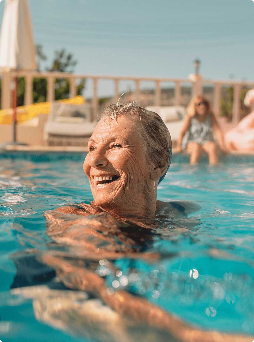 Close-up of an older woman swimming in a sunlit backyard pool, smiling as she enjoys the water.