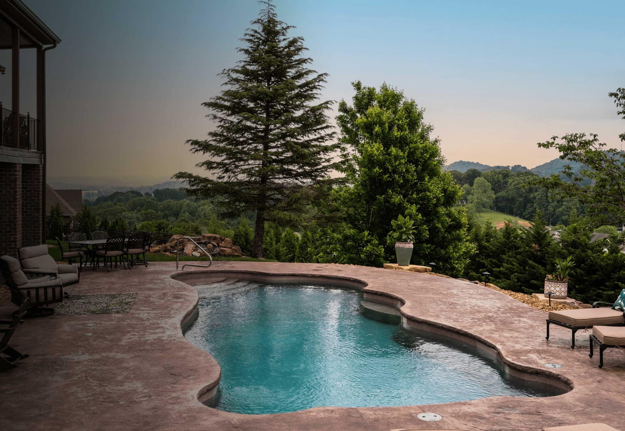 Inground pool in a residential backyard in the mountains, featuring trees and stone.