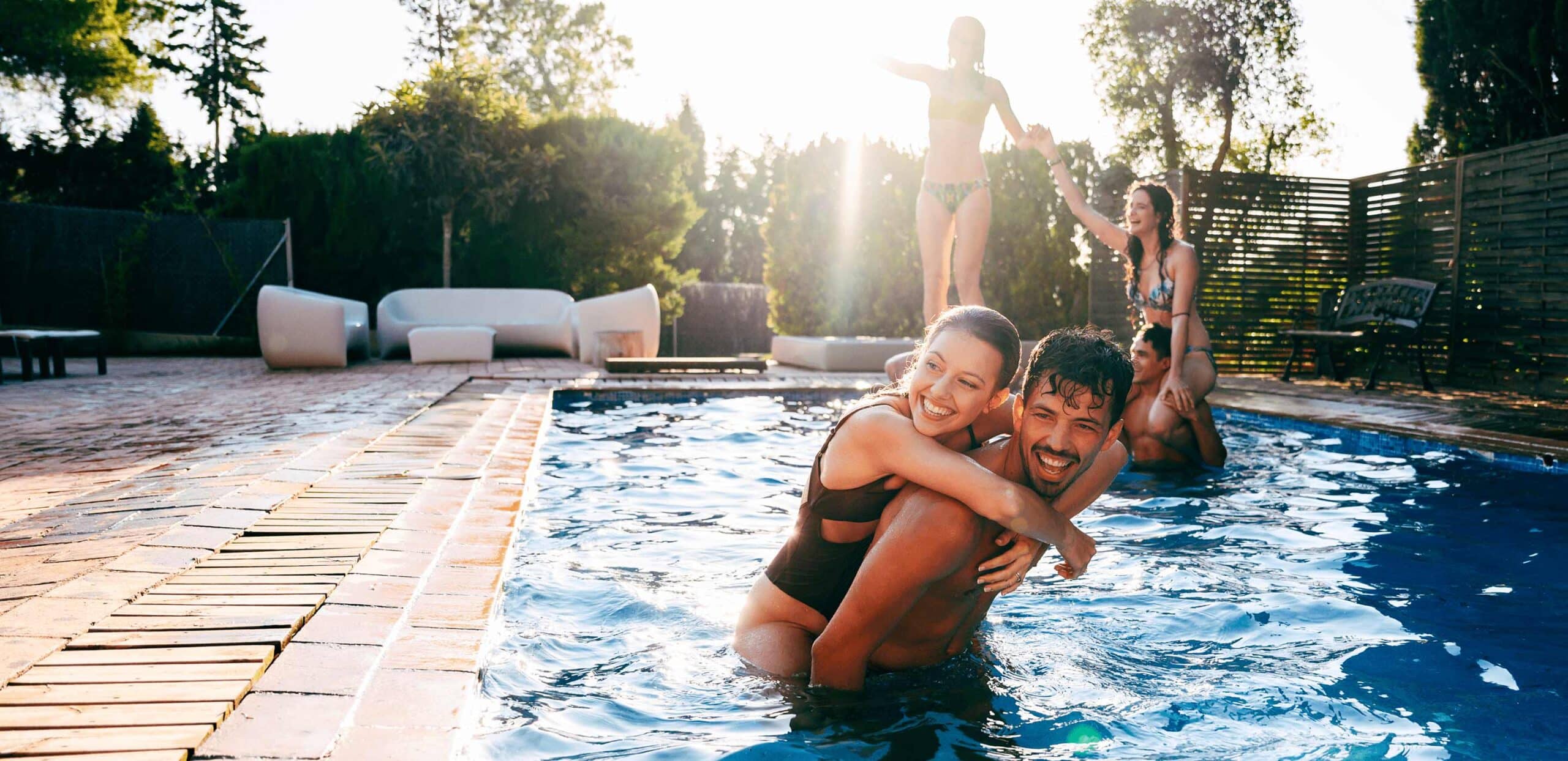 Man holding a child while playing in a pool, both splashing and smiling under evening sun.